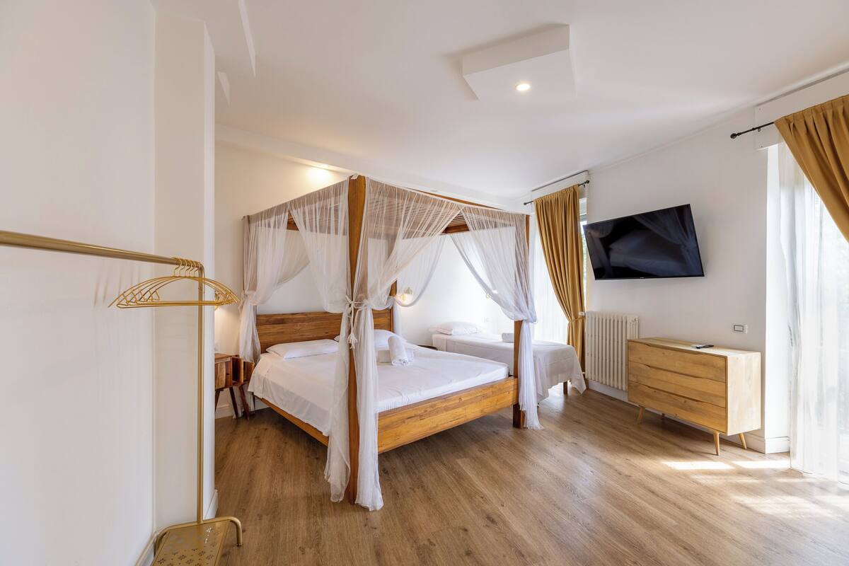 A four-poster bed with a canopy, a wooden dresser, a television mounted on the wall, and a bedside table with a lamp.