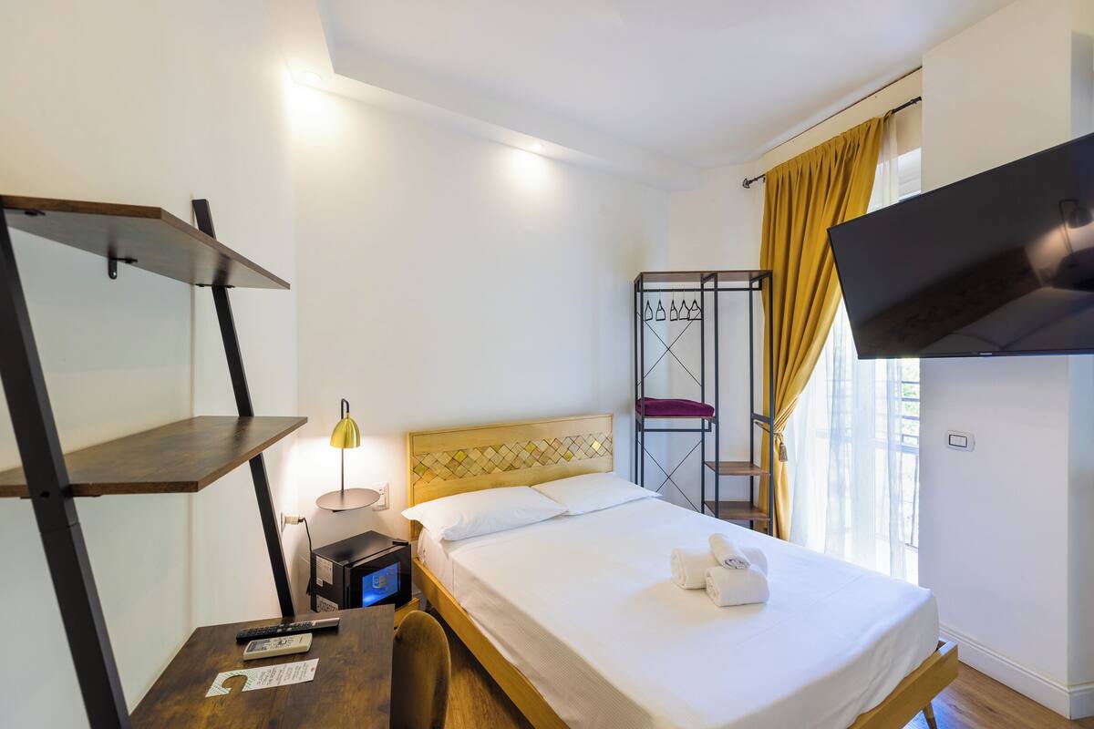 A hotel room with a bed, a desk with a lamp, a TV mounted on the wall, and a window with curtains.