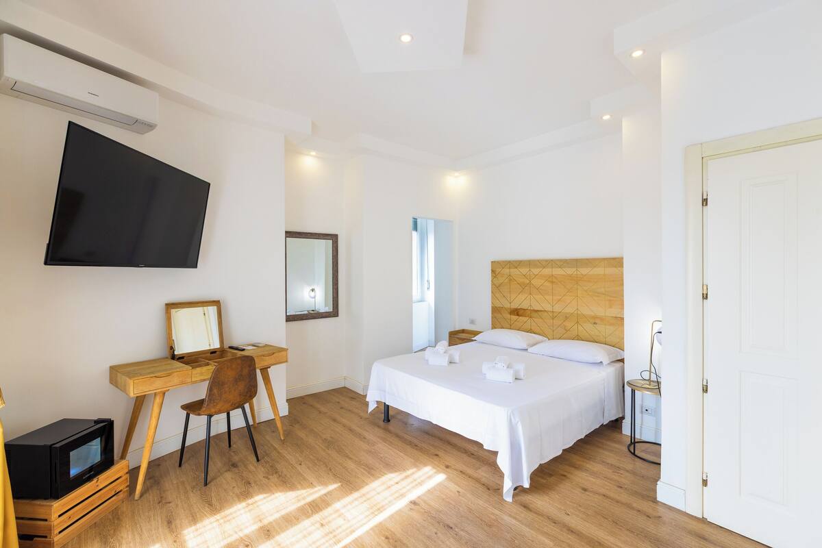 A modern hotel room with a bed, a desk with a chair, a flat-screen TV, and a wooden headboard.