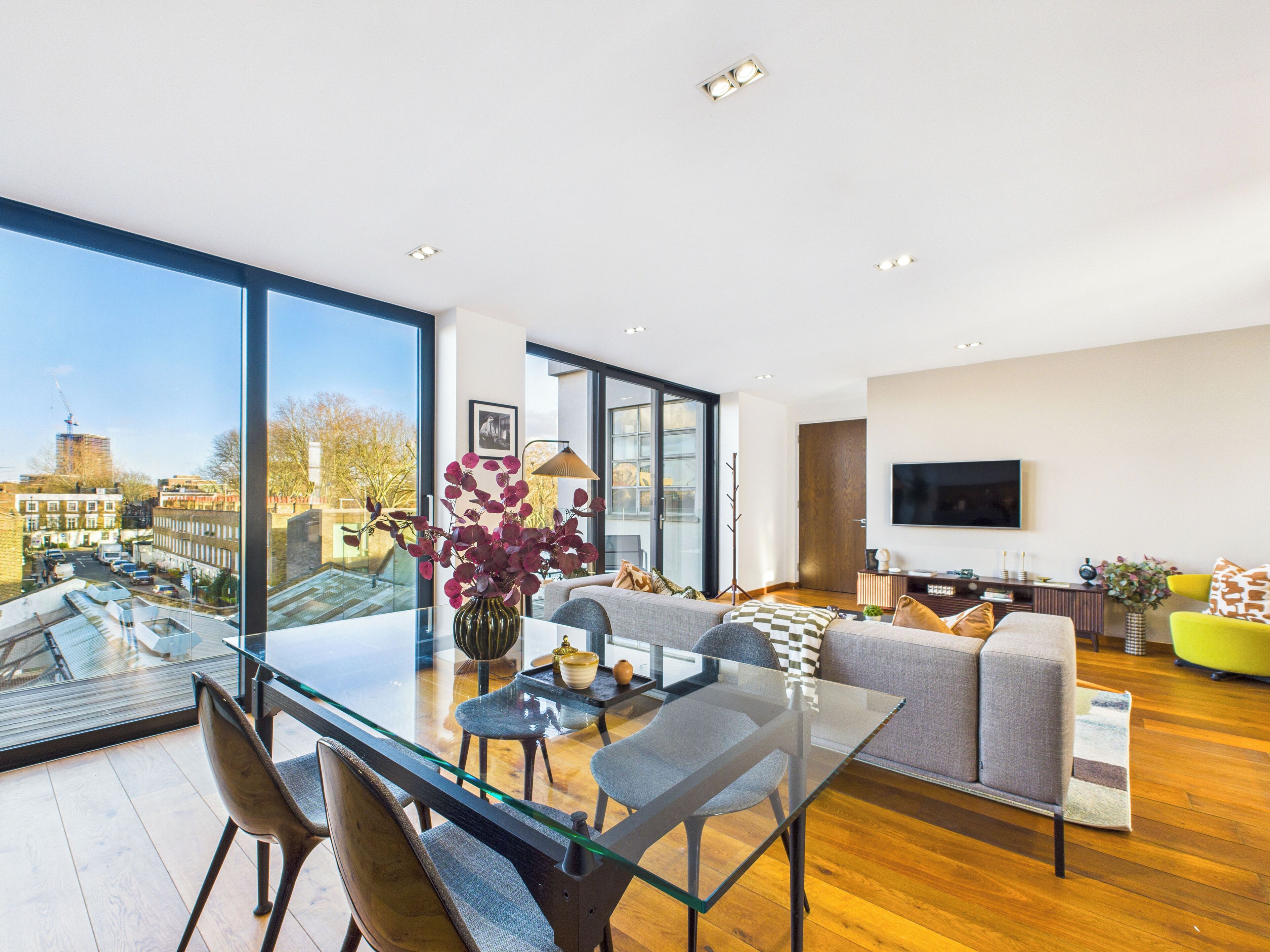 A modern living room with a glass dining table, grey sofa, and a large window offering a city view.