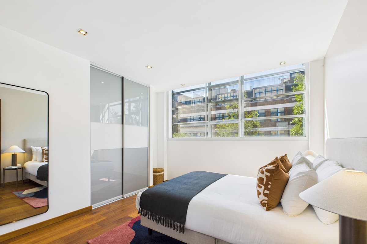 Un dormitorio moderno con una cama grande, un espejo, un ventanal con vistas a la ciudad y suelo de madera.