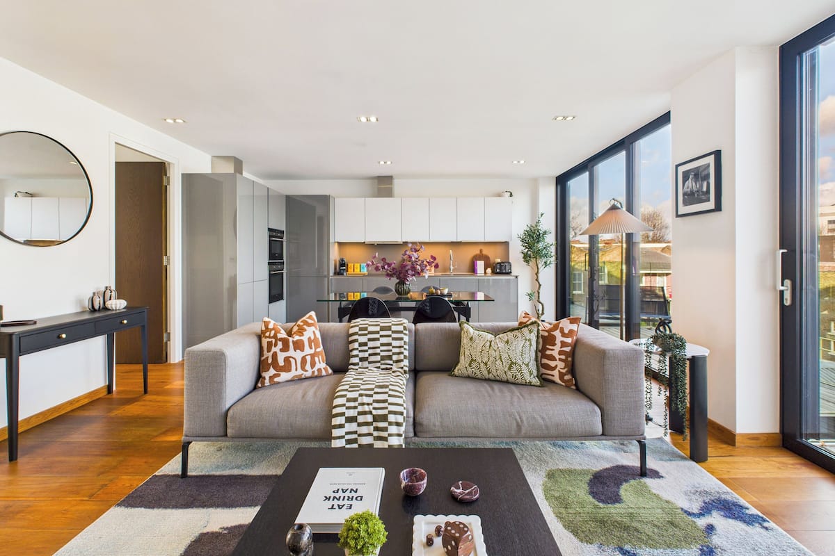 Un salón moderno con un sofá gris, una mesa de centro con un libro, y vistas a la cocina y al exterior.