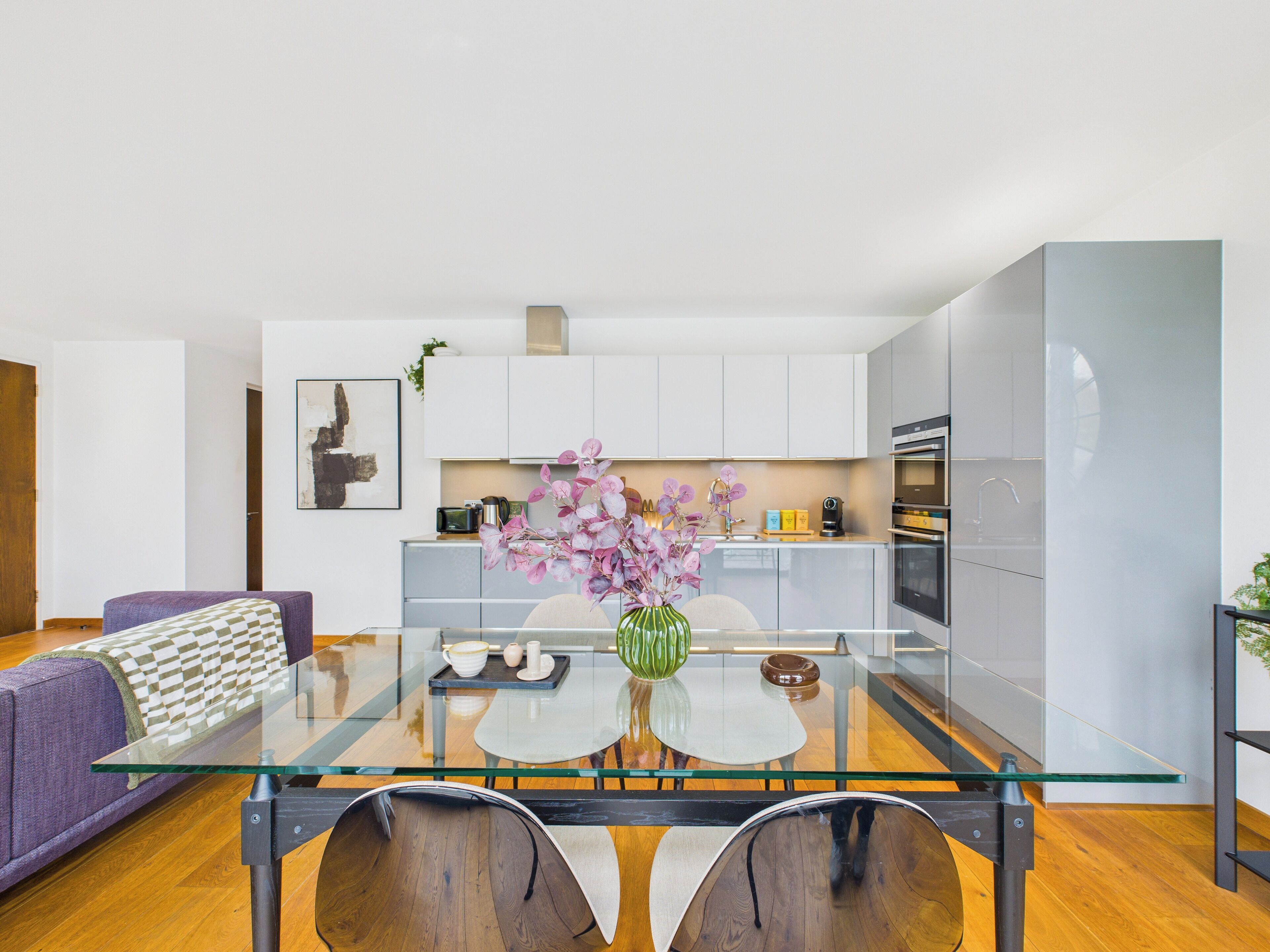 A modern kitchen with white cabinets, a glass dining table, and a purple sofa.