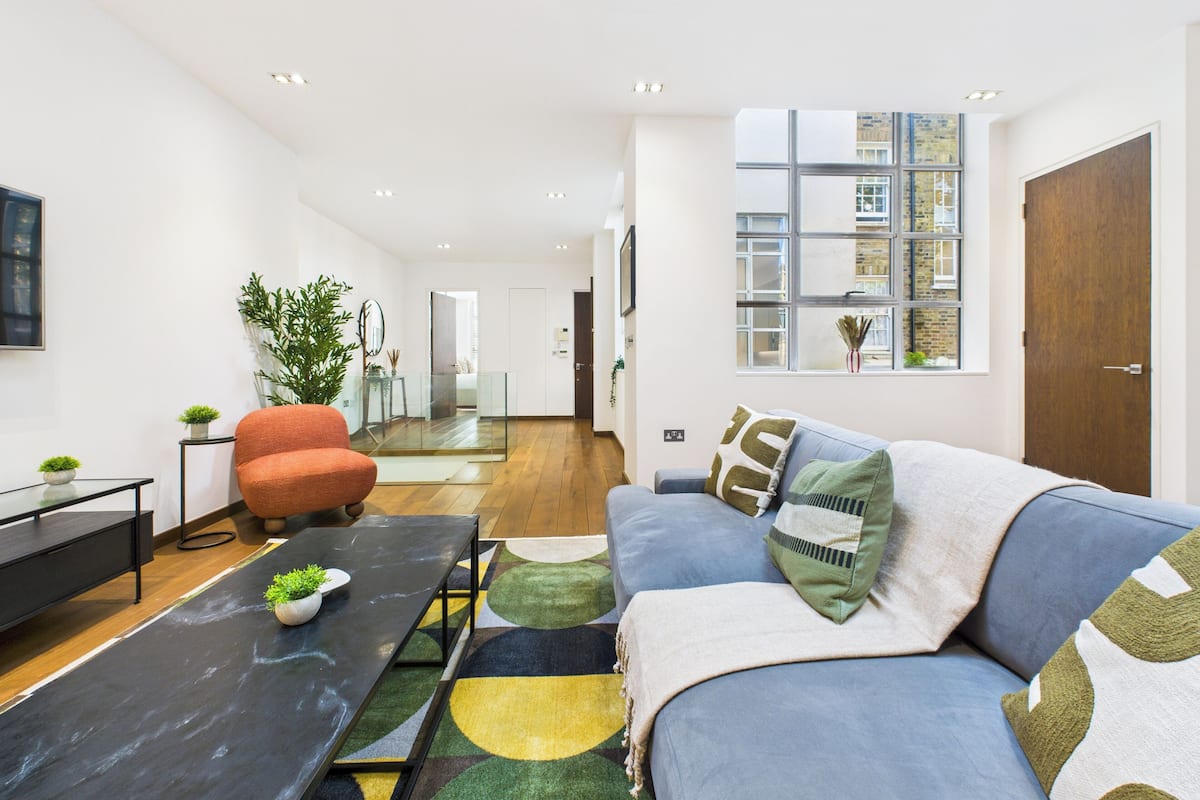 Una sala de estar moderna con un sofá, un sillón y una mesa de centro. Se ve una ventana grande con vistas a edificios.