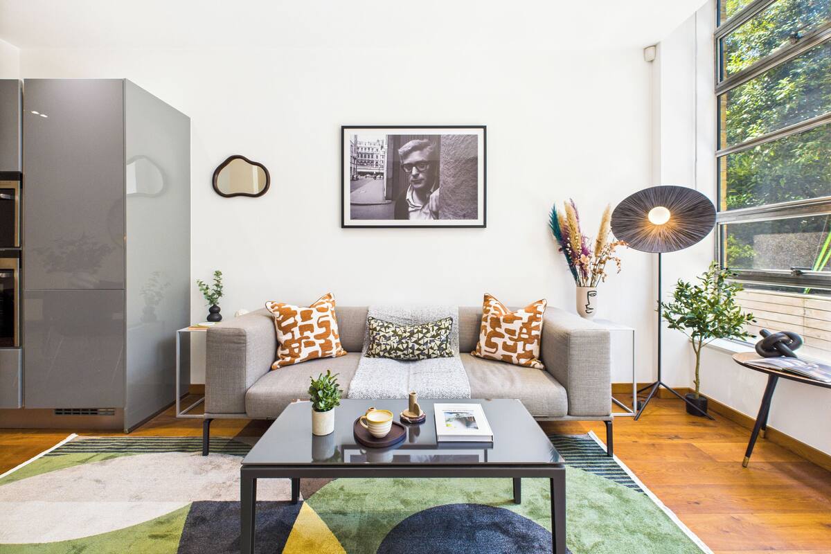 Una sala de estar moderna con un sofá gris, cojines estampados, una mesa de centro con una planta, un cuadro enmarcado en la pared y una lámpara de pie.