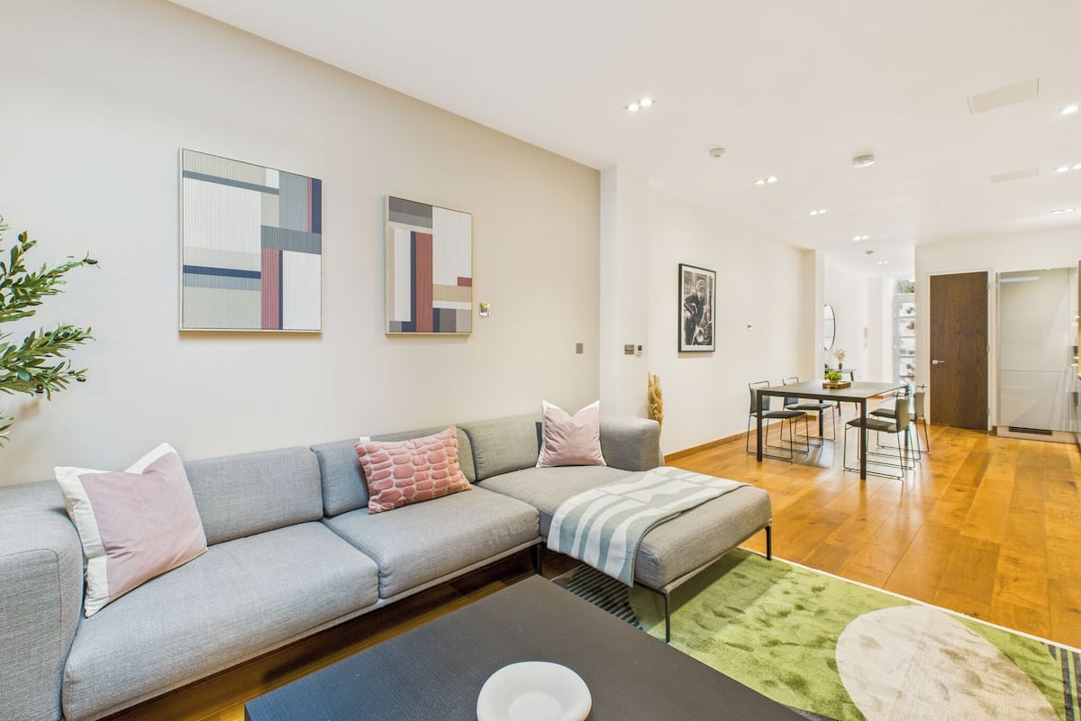 Una sala moderna con un sofá modular gris, un comedor con mesa y sillas, y una cocina al fondo.