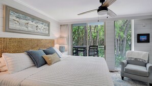 A bedroom with a large bed, a ceiling fan, a view of trees through the window, and a chair by the window. - Villa 407 - Beautiful 2 Bedroom Retreat (Jekyll Island)