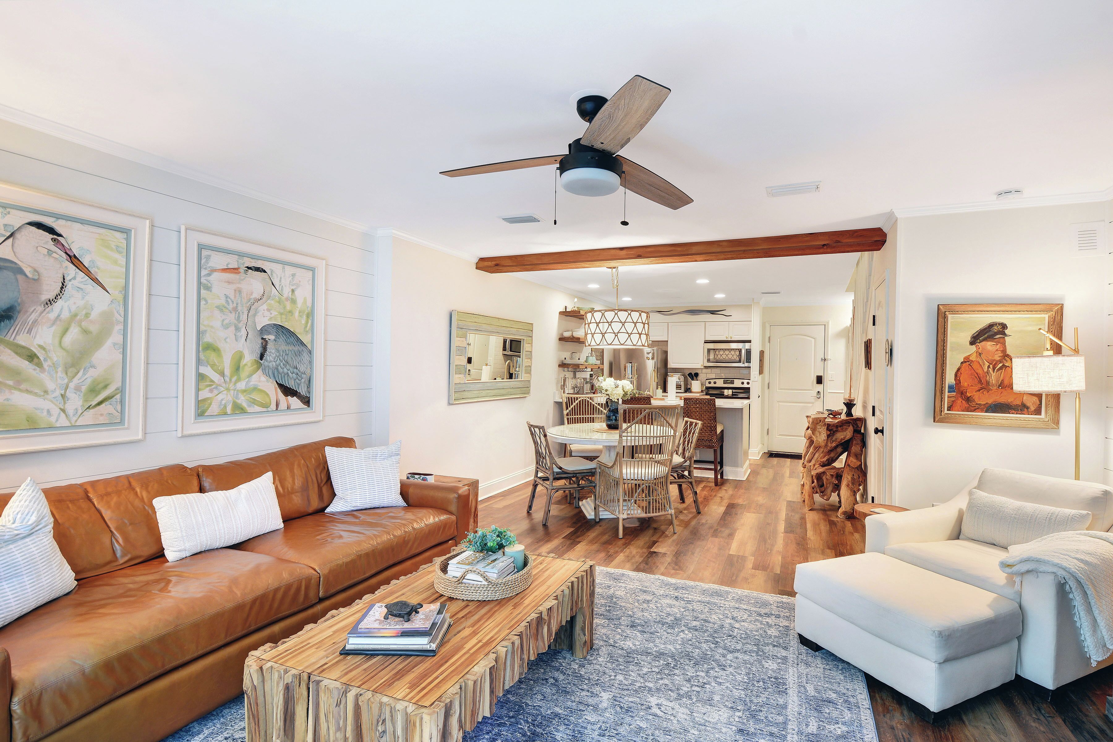 A modern living room with a brown leather sofa, a white armchair, a wooden coffee table, and a dining area with a chandelier.