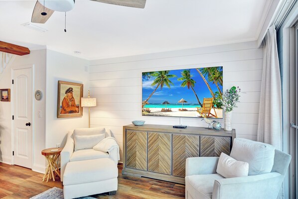A modern living room with a large TV, white sofas, a wooden herringbone cabinet, and a beach-themed painting.