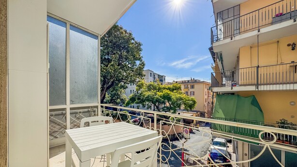 Un balcon avec une table et des chaises, donnant sur une rue bordée de voitures garées et d’immeubles.