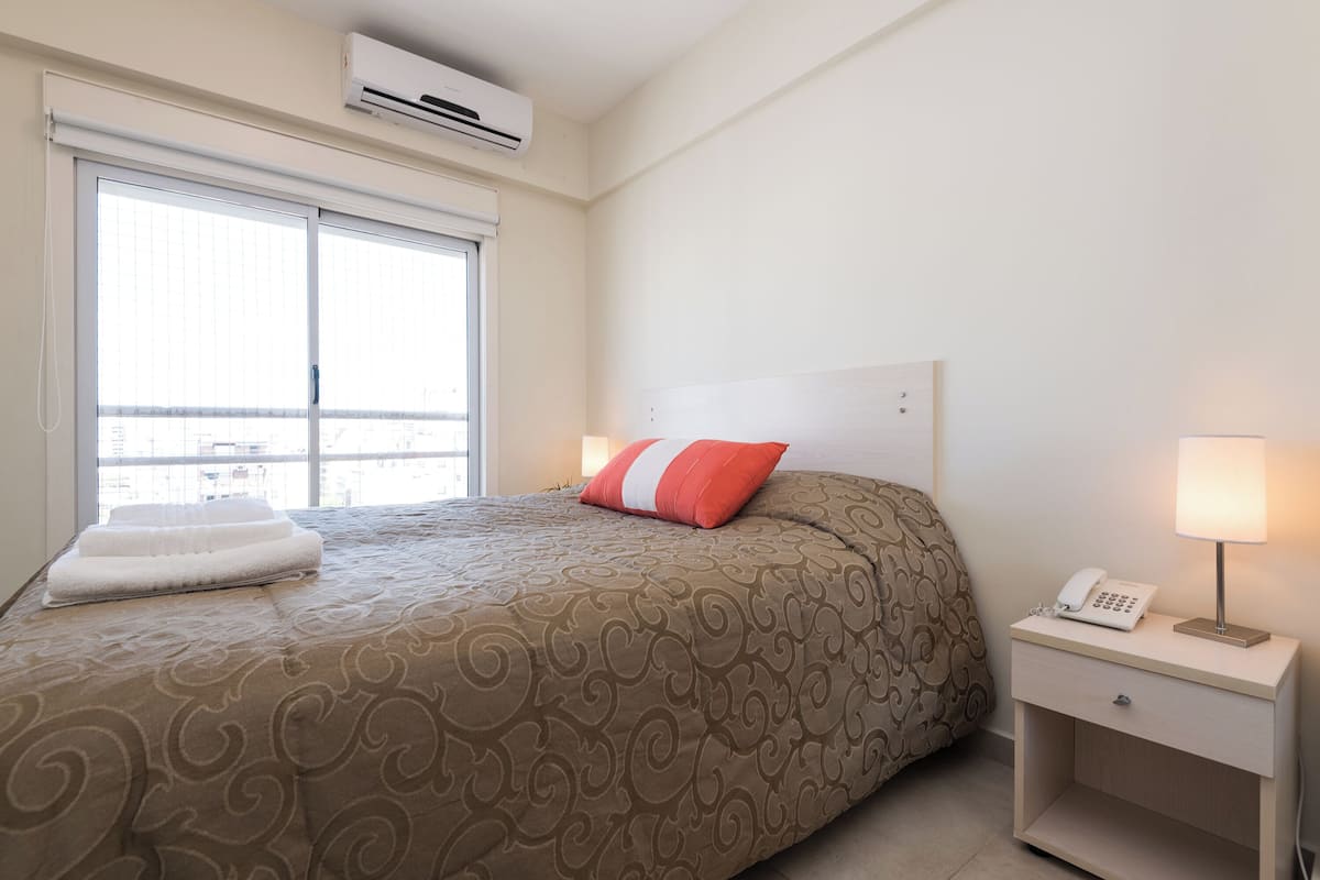 A hotel room with a large bed, a bedside table with a lamp, and a window with blinds.