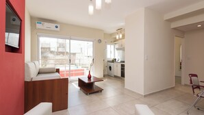 Een moderne woonkamer met een bank, een salontafel en een eethoek. Achtergrond toont een keuken met een wasmachine en een koelkast.