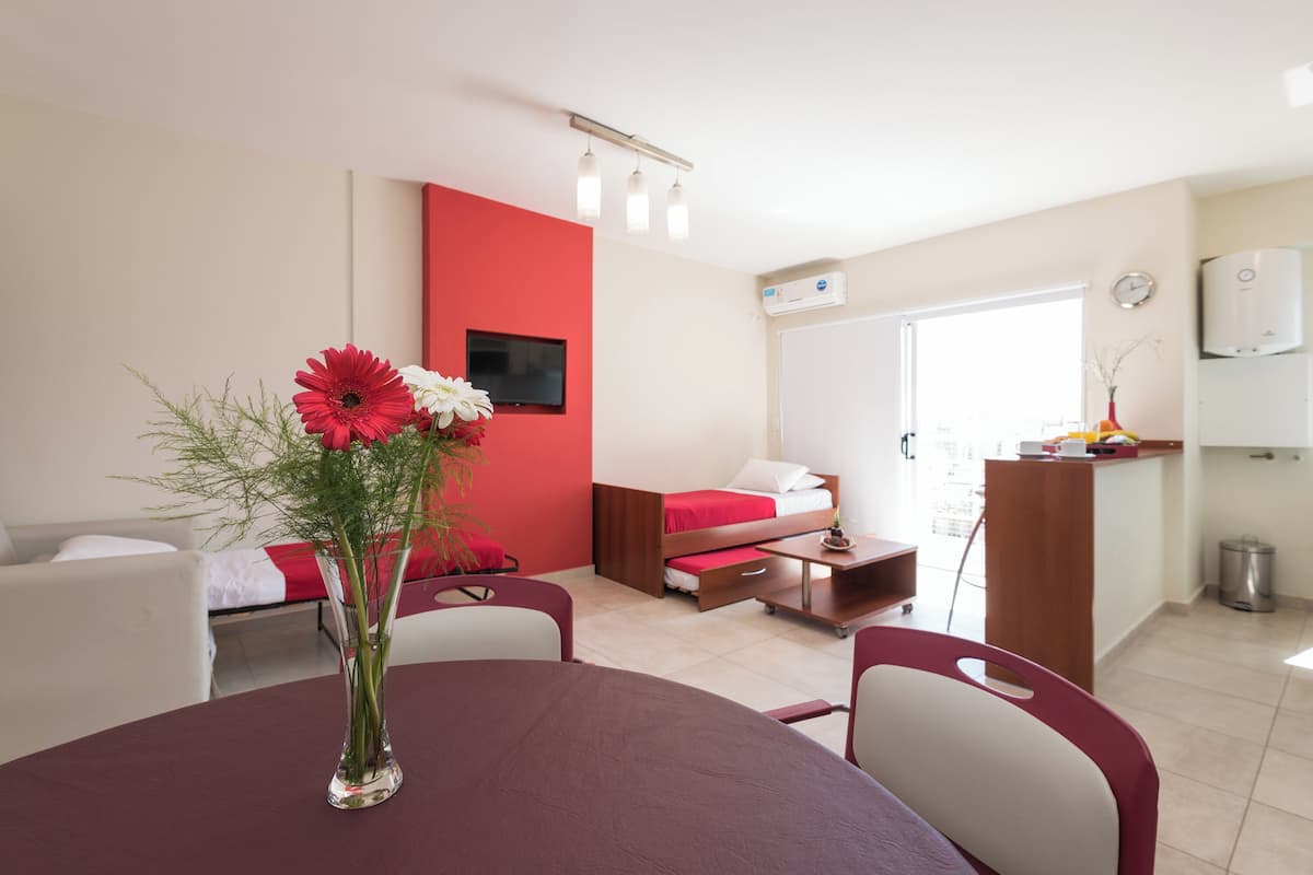 A modern hotel room with a dining area, a red accent wall, a TV, and a kitchenette.