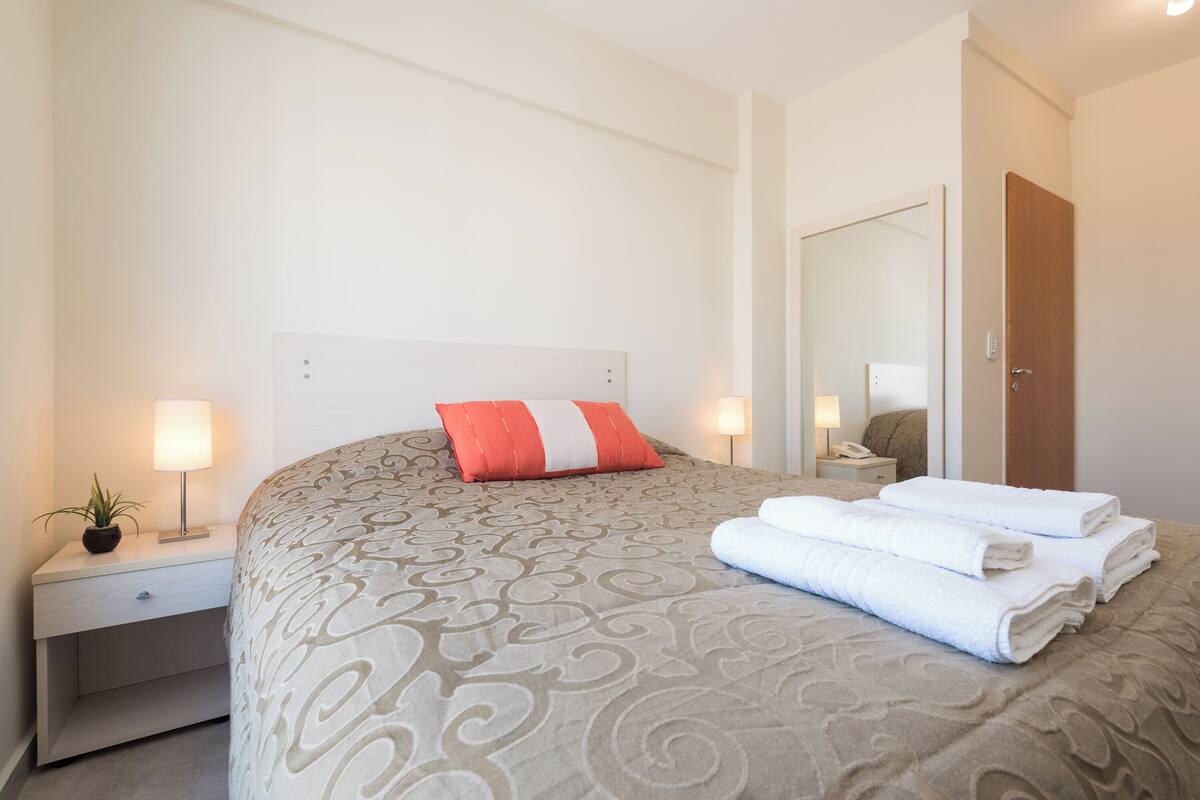 A neatly made bed with a patterned bedspread, a red and white pillow, and folded white towels on the side.