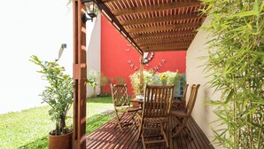 Een houten terras met een rode muur op de achtergrond, een tafel met stoelen en een bloempot.