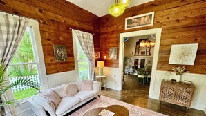 Un salon chaleureux avec un canapé blanc, une table basse ronde et un tapis à motifs. Les murs sont en bois, on aperçoit un coin repas au fond, et on a une vue sur la verdure à l’extérieur.