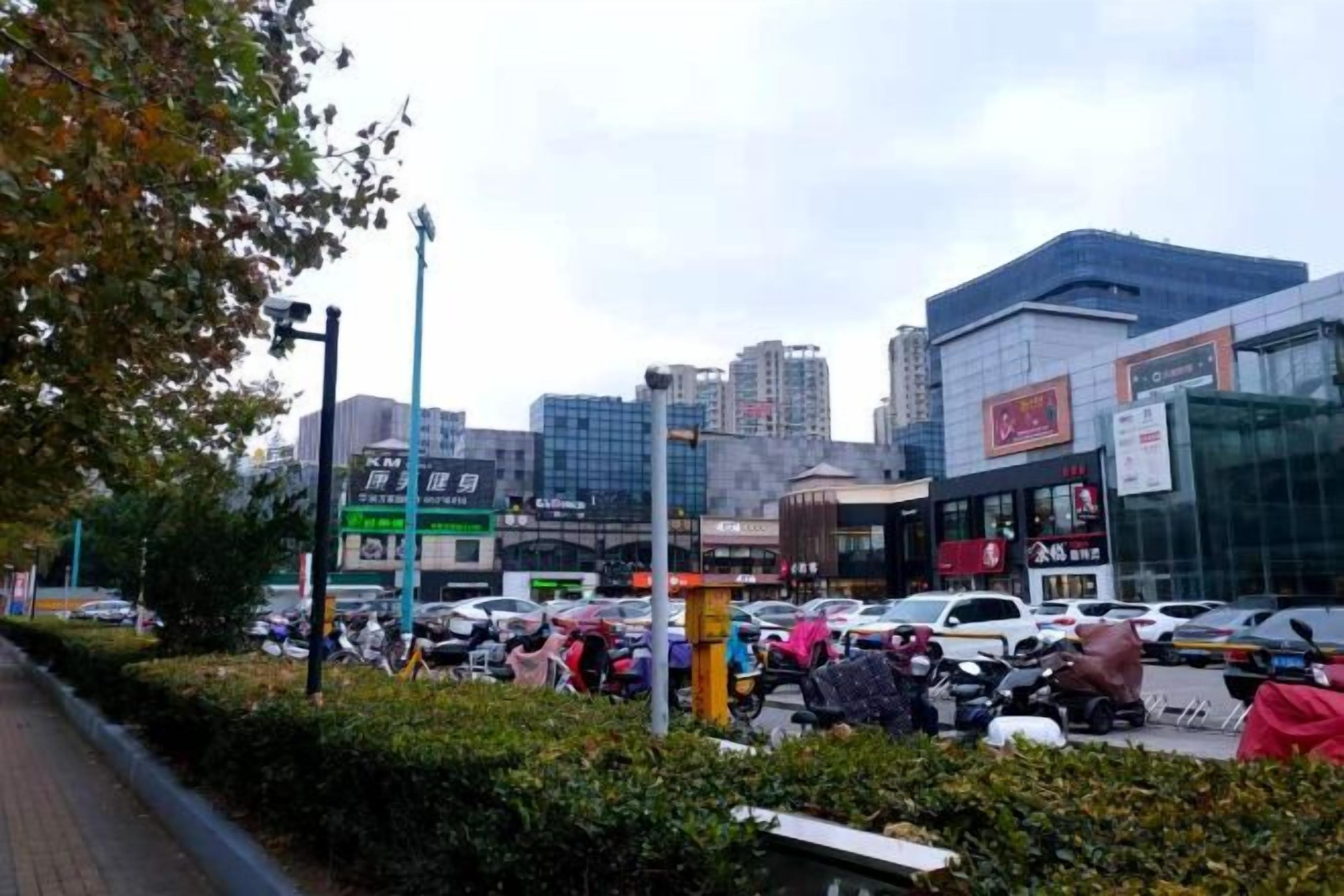 A busy urban street with modern buildings, a parking lot, and a pedestrian walkway.