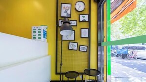 Esquina de una habitación con pared amarilla, un reloj con marco negro y obras de arte enmarcadas. Hay dos sillas negras y una lámpara de pie negra.