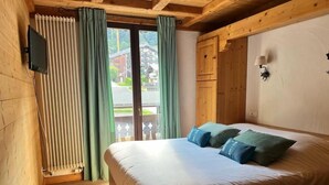 A wooden cabin-style room with a bed, curtains, a radiator, and a view of a mountain and buildings outside. - Fleur Des Neiges  (Morzine)