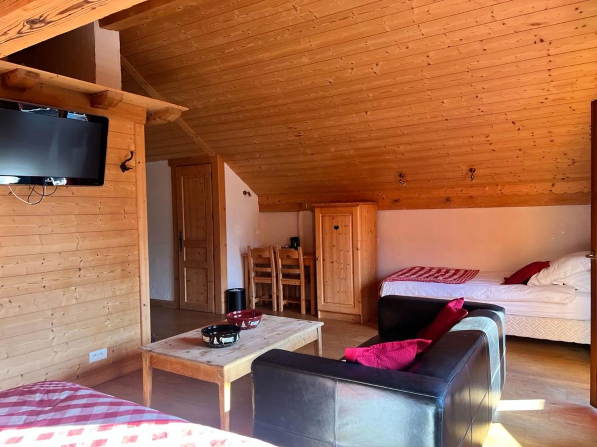 A cozy cabin with a wooden interior, a television mounted on the wall, a dining table, a sofa, and a bed with a red and white checkered blanket.