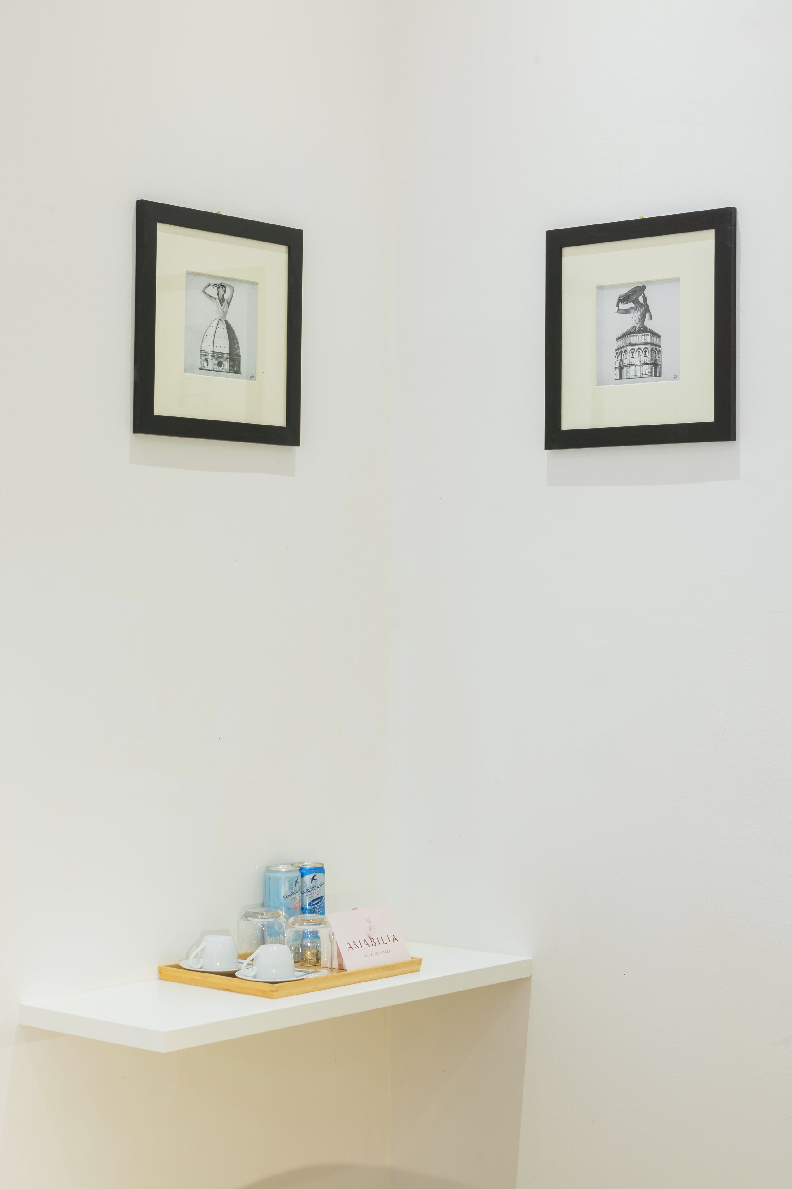 Dos obras de arte blanco y negro enmarcadas sobre una pared blanca, una pequeña mesa blanca con una bandeja que sostiene una lata, un vaso y una taza, y un letrero con la palabra "AMABILIA".