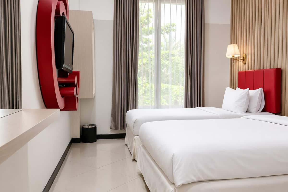 Habitación de hotel con dos camas, un televisor instalado en una pared roja, una ventana con cortinas y una lámpara de pared.
