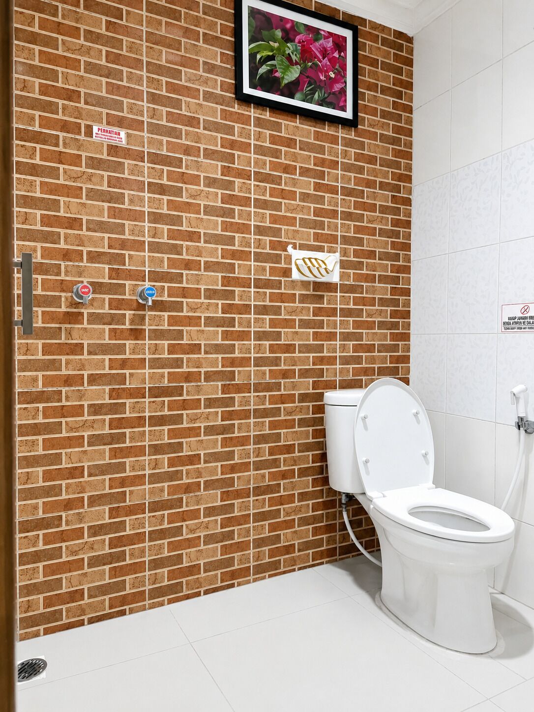 Un baño con pared de ladrillo, un inodoro, una ducha y una imagen de flores.