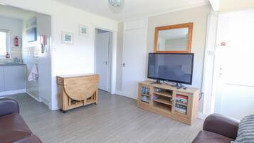 Una sala de estar con televisión, muebles de madera y una cocina al fondo.