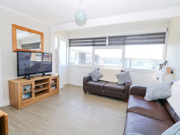 A modern living room with a flat-screen TV on a wooden stand, a brown leather sofa, a wooden cabinet with books, and a large window with blinds. - Chalet 39 (Great Yarmouth)
