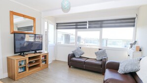 A modern living room with a flat-screen TV on a wooden stand, a brown leather sofa, a wooden cabinet with books, and a large window with blinds. - Chalet 38 (Great Yarmouth)