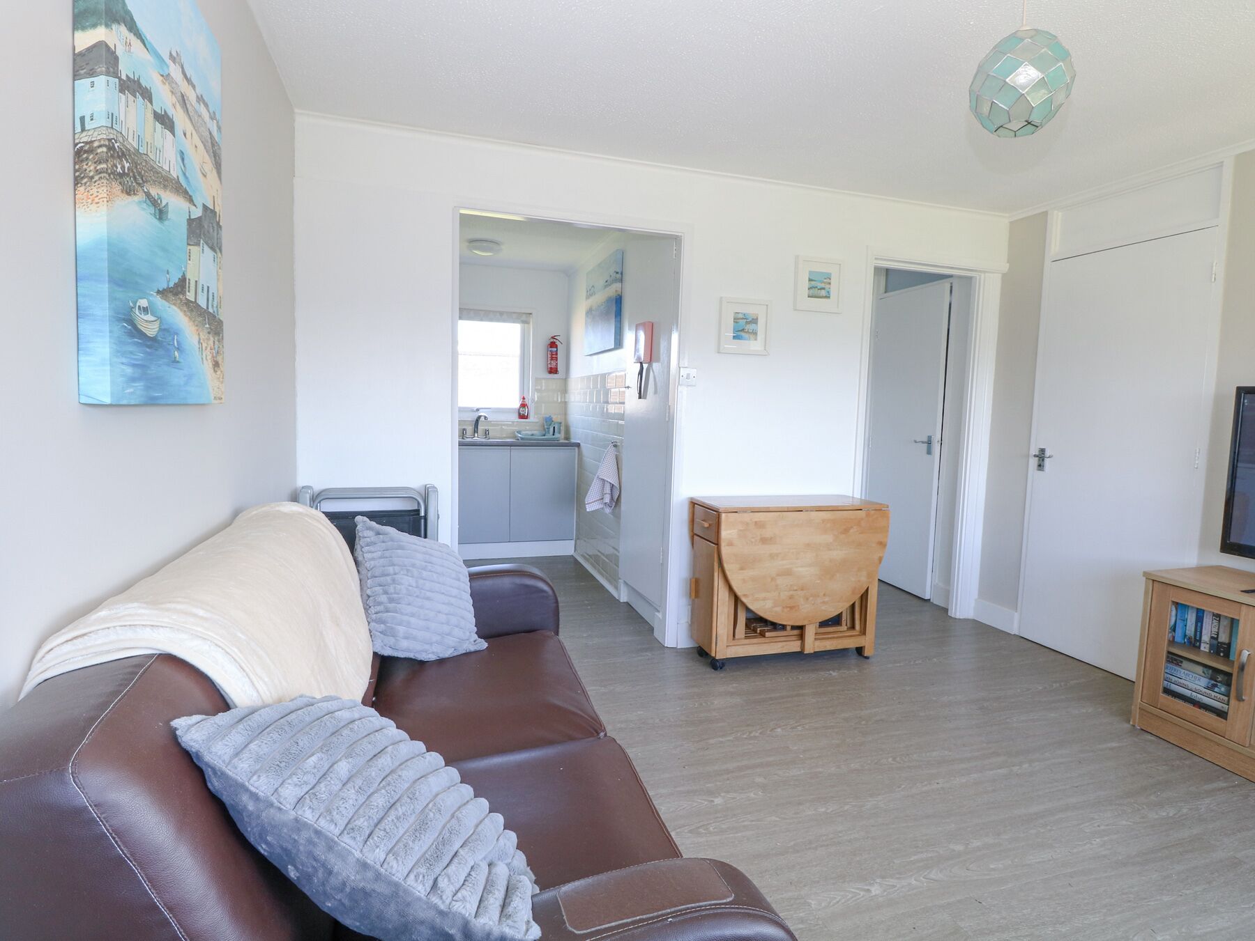 A living room with a brown leather sofa, a wooden side table, and a painting of a coastal scene. A kitchen is visible in the background.