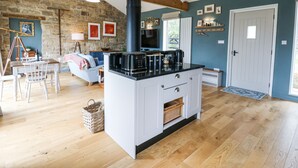 A cozy living room with a stone wall, wooden beams, a black stove, and a dining area with a table and chairs. - The Barn (Riding Mill)