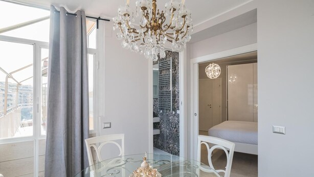 A modern living room with a glass dining table, white chairs, a chandelier, and a balcony with a view of the city. - B&B Damagi (Rome)