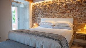 Un dormitorio con pared de piedra, una cama con ropa blanca, un banco y una mesita de noche con lámpara.