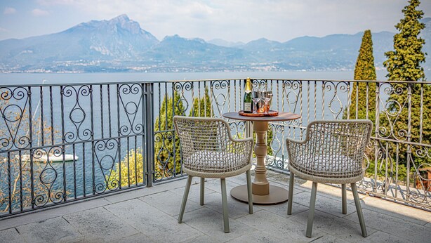 Un balcón con dos sillas de mimbre y una mesa, con vistas a un paisaje montañoso y un cuerpo de agua.