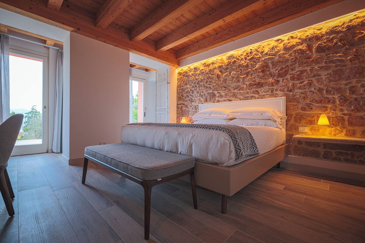 A bedroom with a stone wall, a bed with white bedding, a bedside table with a lamp, and a bench.