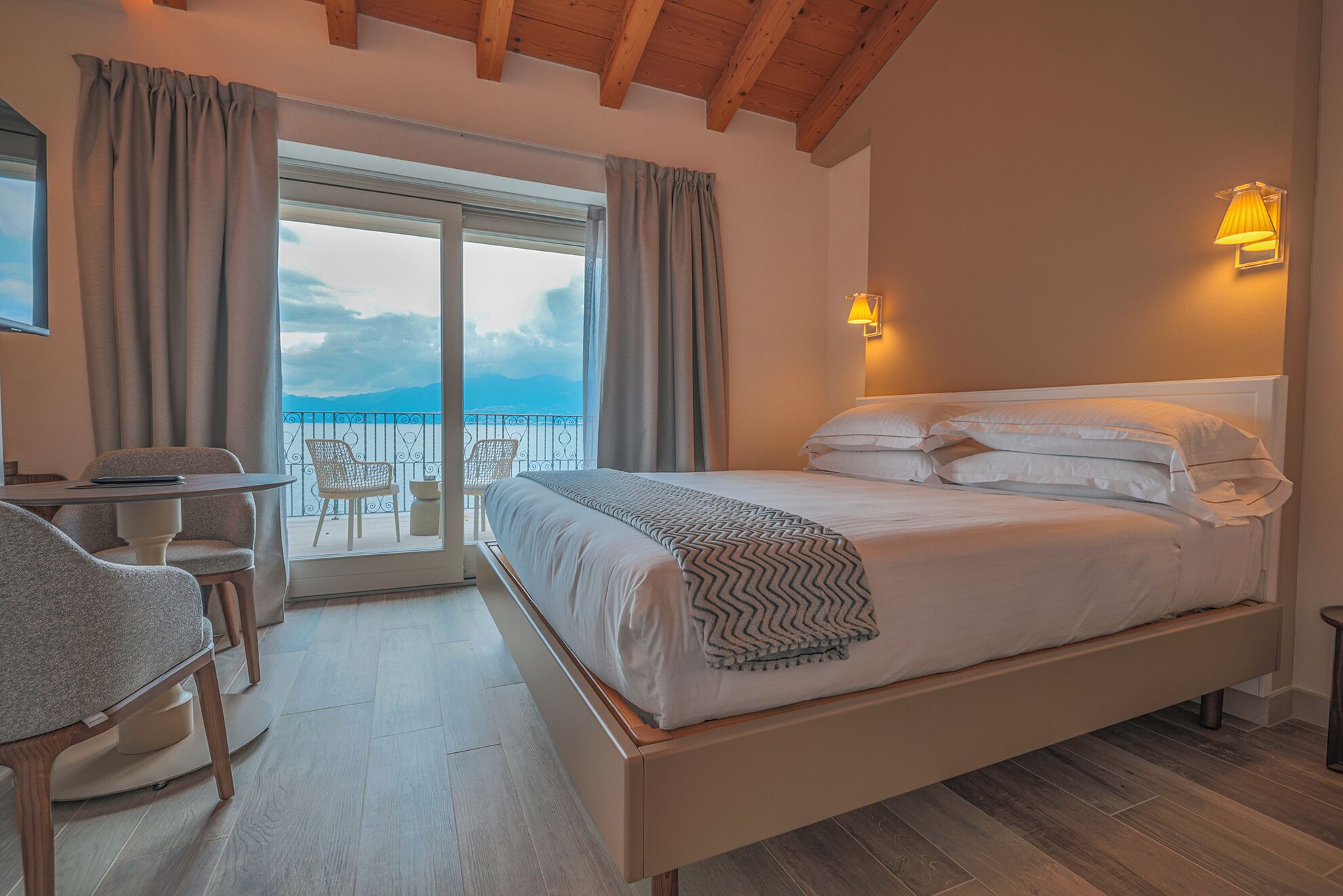 Un dormitorio con cama, escritorio y silla. Tiene un balcón con vistas al cielo y a las montañas.