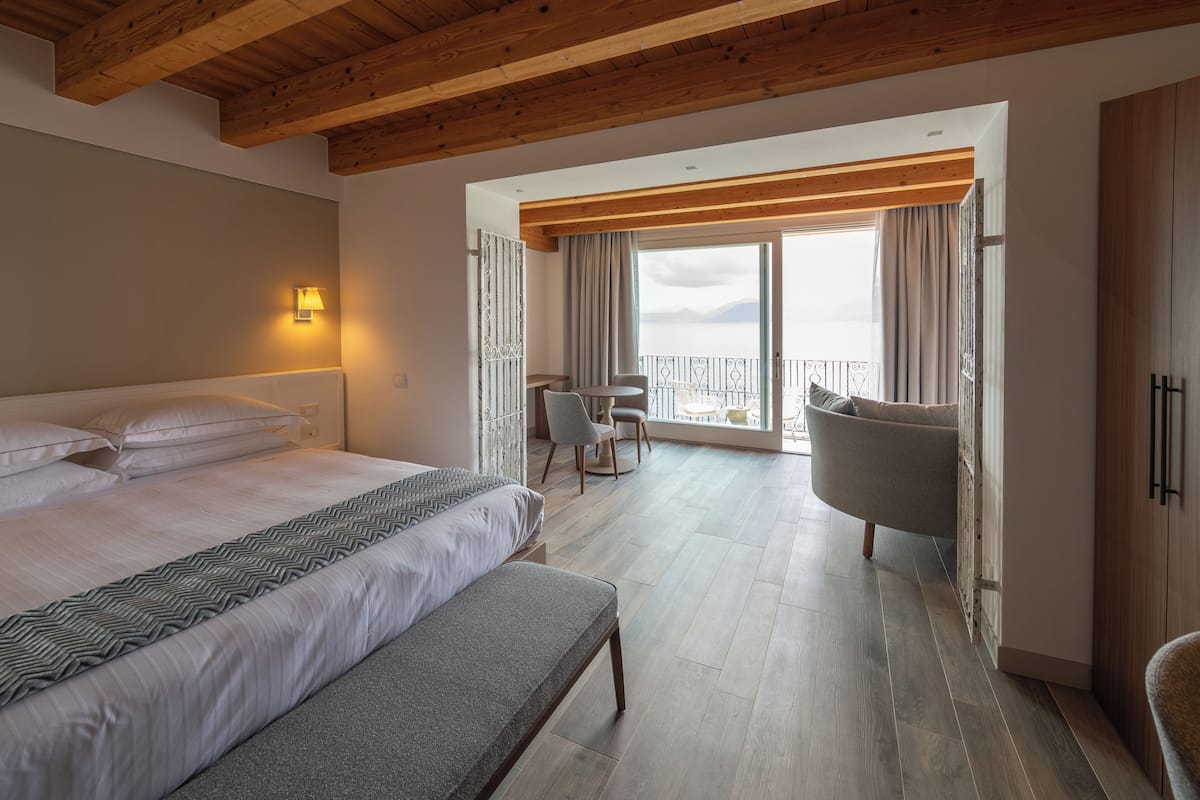 A hotel room with a large bed, a bench, a dining table with chairs, and a balcony with a view.