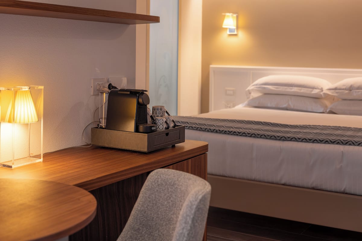 A hotel room with a wooden desk, a lamp, a coffee maker, and a bed with pillows.