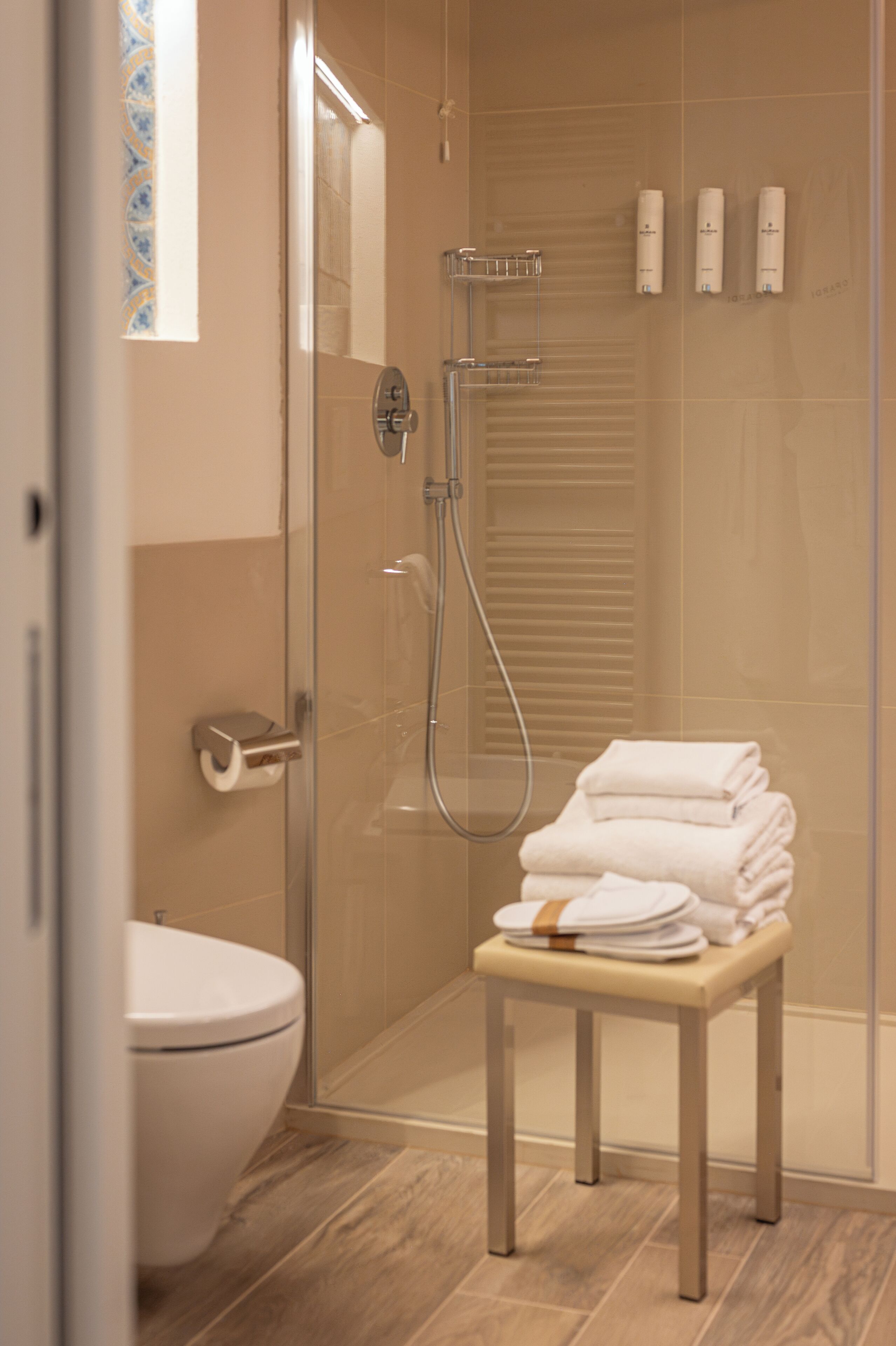 A modern bathroom with a glass-enclosed shower, a toilet, and a stack of folded towels on a bench.