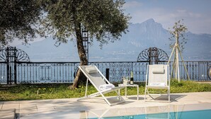 Zona junto a la piscina con dos sillones de descanso, una mesa con una botella y vasos, y vista a una montaña y un cuerpo de agua.