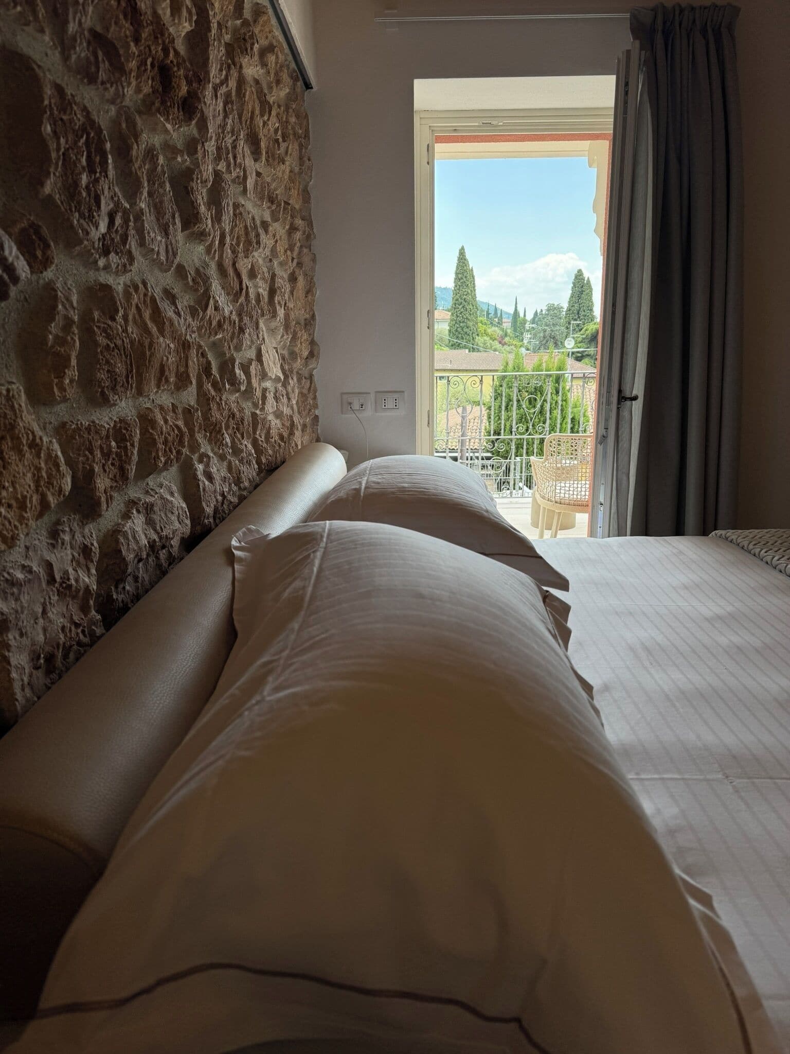 Un dormitorio con pared de piedra, cama con ropa blanca y balcón con vistas a la vegetación.