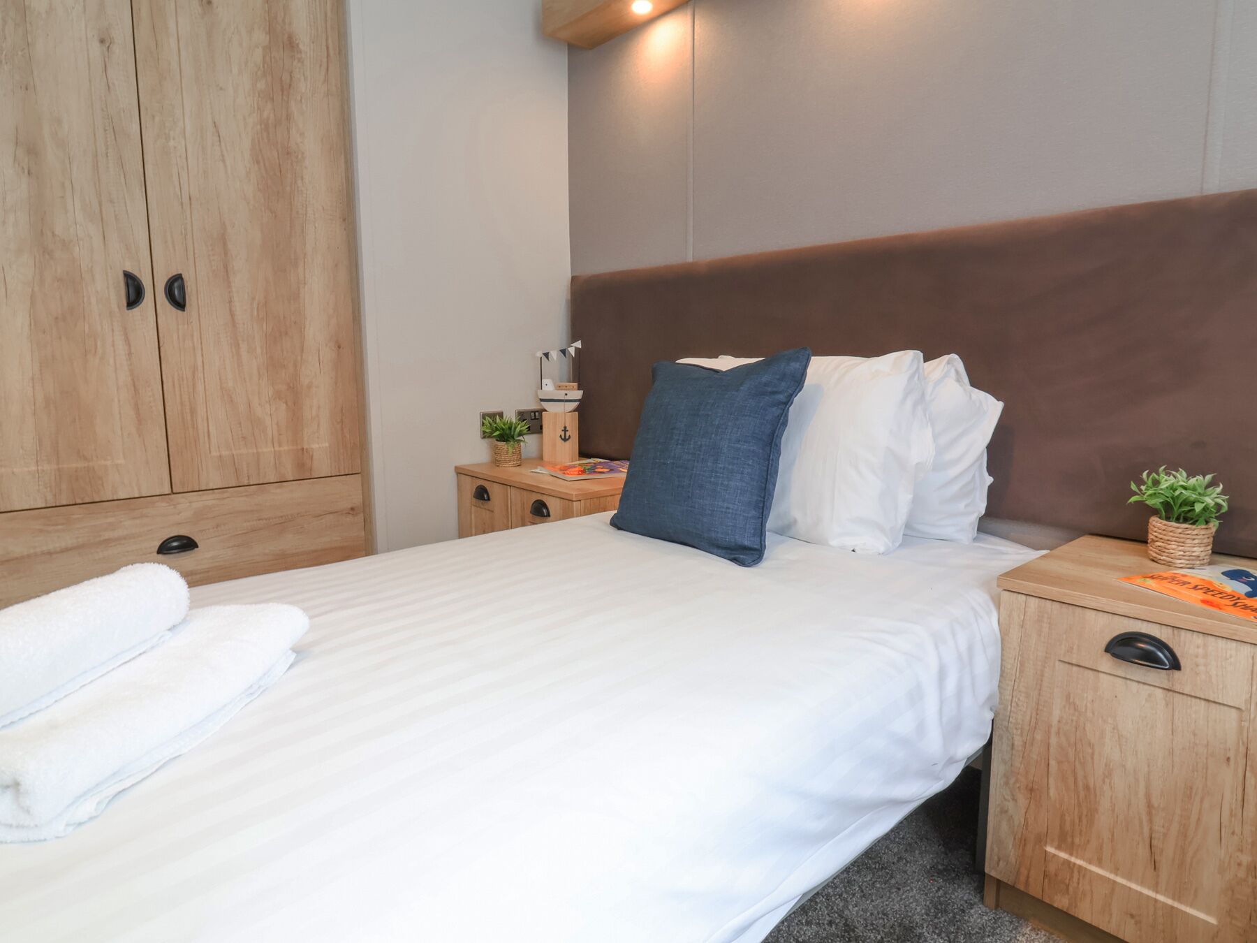A neatly made bed with white linens, a blue pillow, and a white pillow. There is a wooden nightstand with a small plant and a decorative item. A wooden wardrobe is visible in the background.