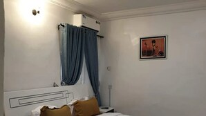 Egy szállodai szoba, amelyben található egy ágy, egy éjjeliszemély, egy lámpa, egy kép a falon, és egy ablak, amelyen függöny van.