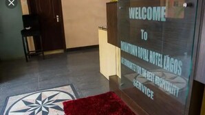Egy fogadótér, amelyen a „Üdvözöljük a Downtown Royal Hotel Lagosban!” feliratú tábla, egy piros szőnyeg és egy mintás padlóburkolat található.