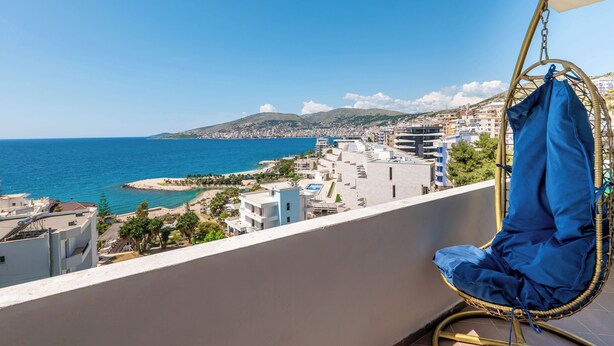 Un balcón con una silla colgante que da a una vista de la ciudad costera, con edificios y una playa.