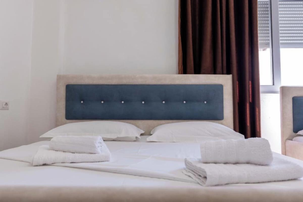 A neatly made bed with white linens and towels, a tufted headboard, and a window with curtains.
