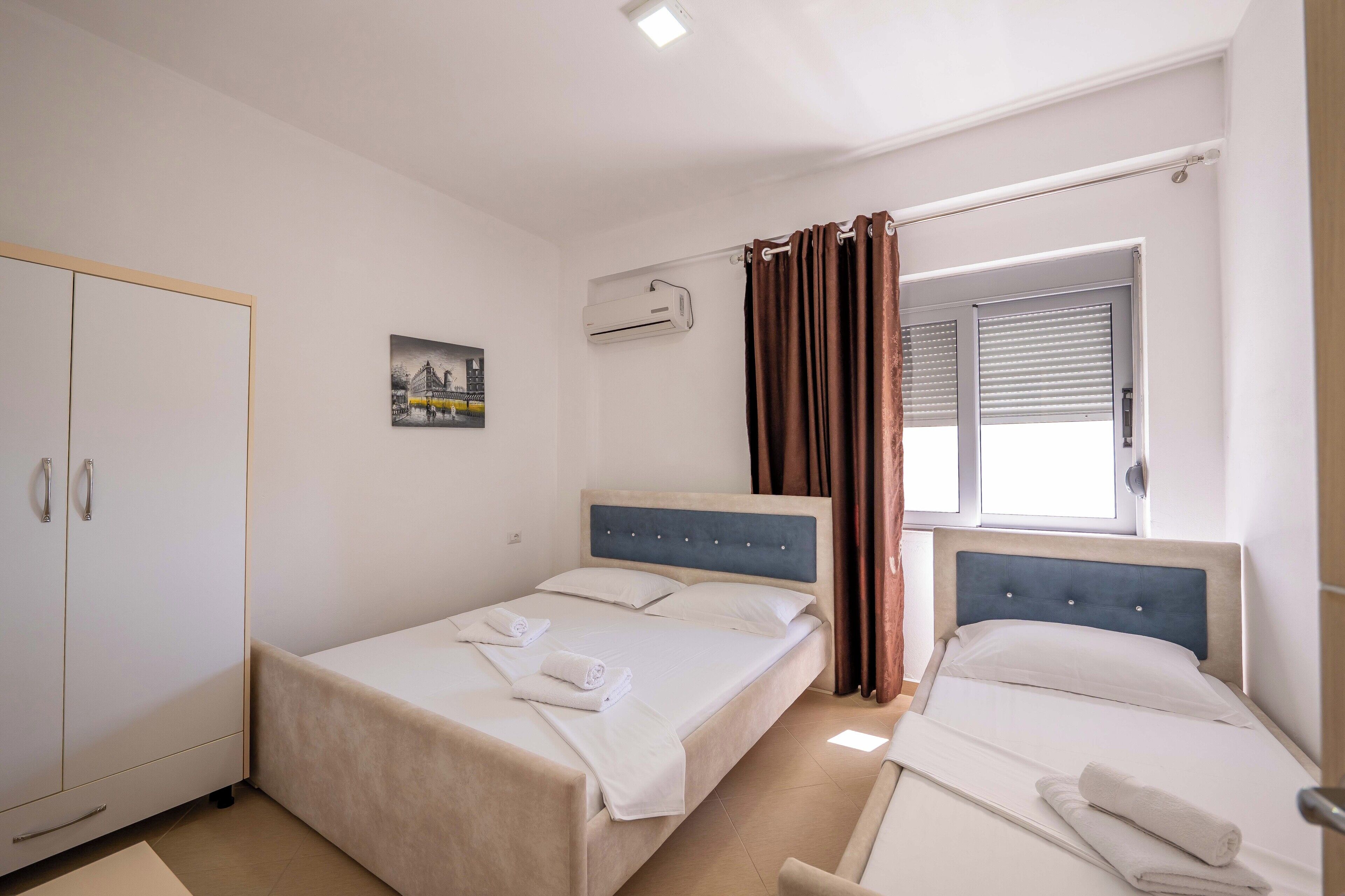 A hotel room with two beds, a wardrobe, an air conditioning unit, and a window with curtains.