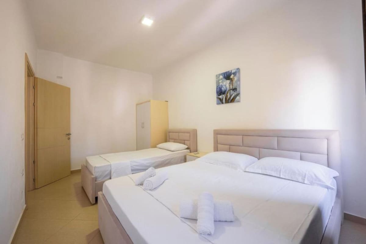 A hotel room with two beds, a door, a nightstand, and a painting on the wall.