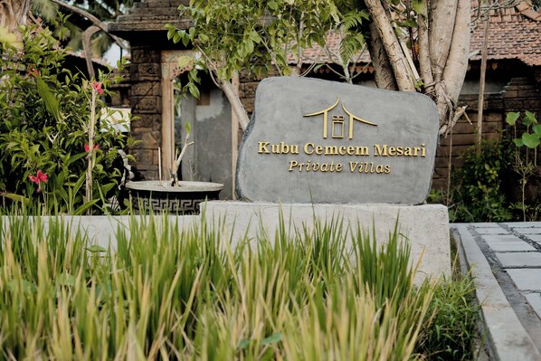 A sign for Kubu Cemcen Mesari Private Villas with a stone base and gold lettering, surrounded by greenery and a stone building in the background. - Kubu Cemcem Mesari Private Villas (Ubud)